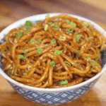 A full bowl of glossy garlic noodles is topped with scallions and black pepper, ready to be served hot.