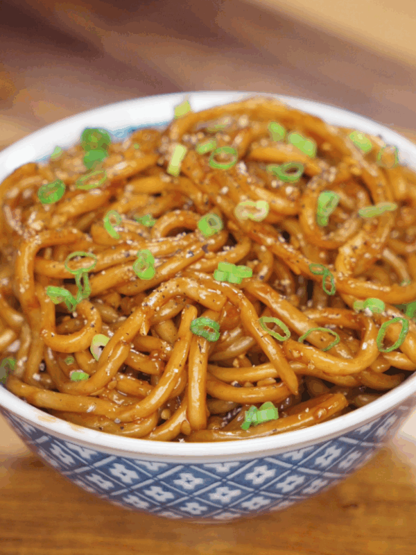 A full bowl of glossy garlic noodles is topped with scallions and black pepper, ready to be served hot.