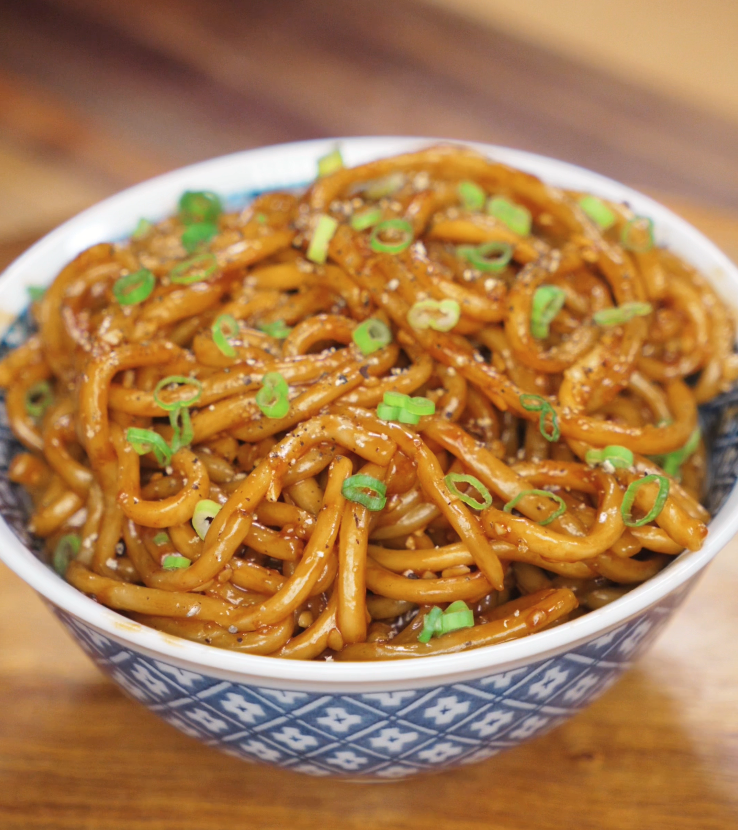 A full bowl of glossy garlic noodles is topped with scallions and black pepper, ready to be served hot.