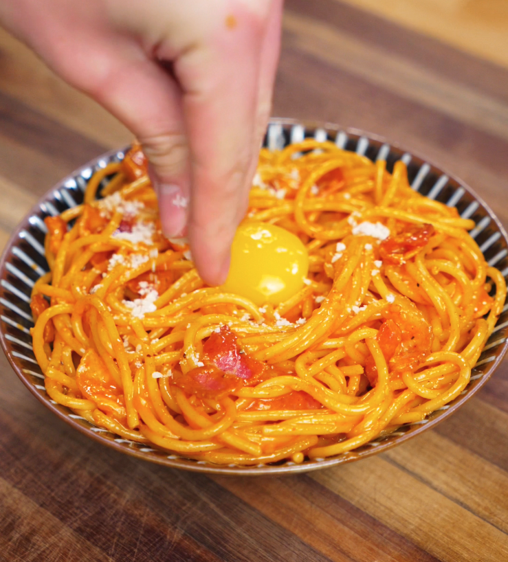 A hand sprinkles parmesan over a bowl of gochujang carbonara, finished with a raw egg yolk nestled in the center for extra richness.