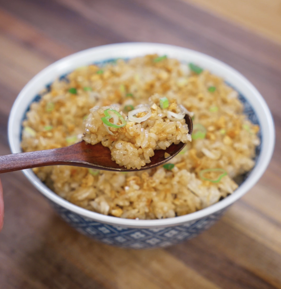 A wooden spoon lifts a glossy bite of garlic fried rice, topped with scallions and crispy golden garlic, revealing its rich texture and color.