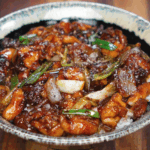 A ceramic bowl filled with saucy black pepper chicken, glossy onions, scallions, and tender chicken thighs served over white rice.