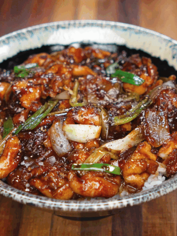 A ceramic bowl filled with saucy black pepper chicken, glossy onions, scallions, and tender chicken thighs served over white rice.