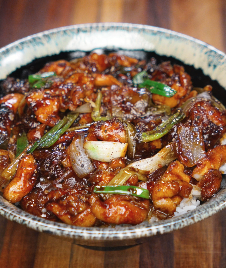 A ceramic bowl filled with saucy black pepper chicken, glossy onions, scallions, and tender chicken thighs served over white rice.