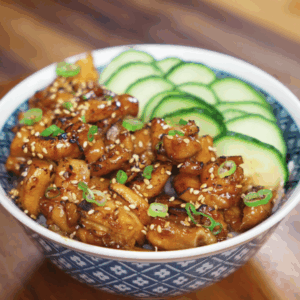 A full bowl of garlic butter chicken with glossy, caramelized chicken chunks, sesame seeds, scallions, and thin cucumber slices over a bed of white rice.