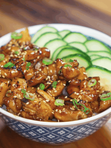 A full bowl of garlic butter chicken with glossy, caramelized chicken chunks, sesame seeds, scallions, and thin cucumber slices over a bed of white rice.