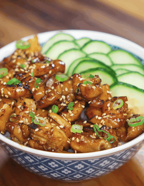 A full bowl of garlic butter chicken with glossy, caramelized chicken chunks, sesame seeds, scallions, and thin cucumber slices over a bed of white rice.