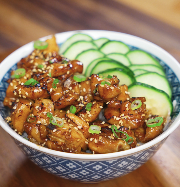 A full bowl of garlic butter chicken with glossy, caramelized chicken chunks, sesame seeds, scallions, and thin cucumber slices over a bed of white rice.