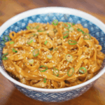 A full bowl of glossy sesame peanut noodles topped with sliced scallions and sesame seeds, the sauce clinging to every strand with a rich sheen.