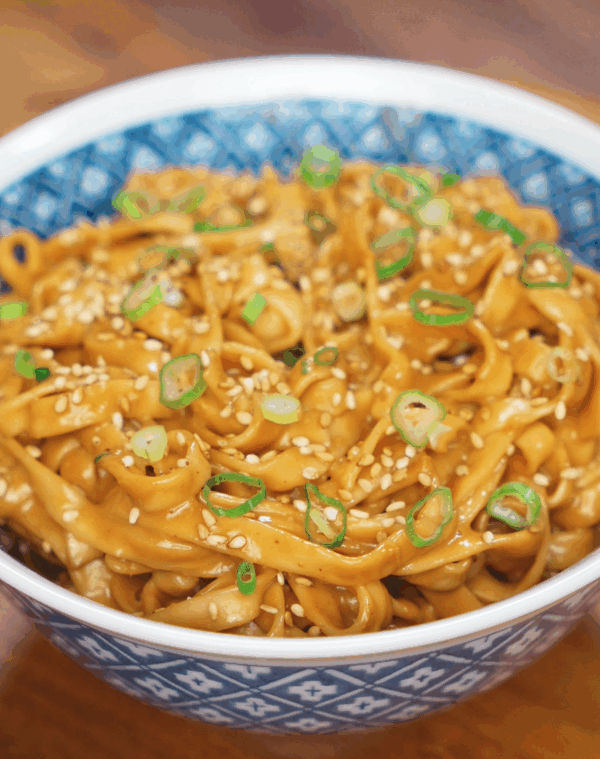 A full bowl of glossy sesame peanut noodles topped with sliced scallions and sesame seeds, the sauce clinging to every strand with a rich sheen.