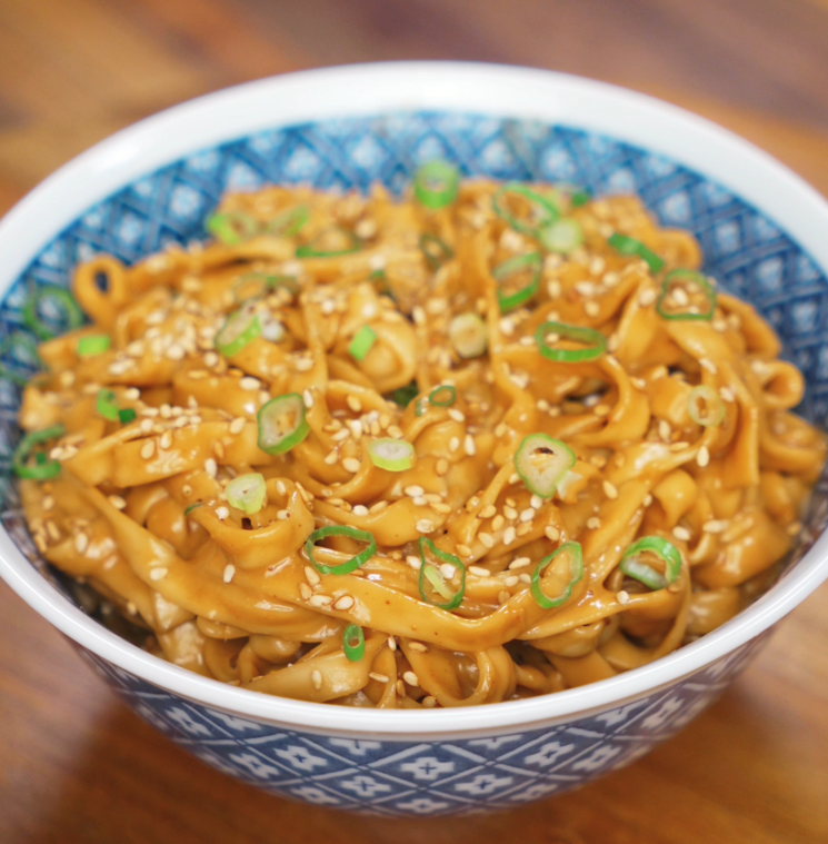 A full bowl of glossy sesame peanut noodles topped with sliced scallions and sesame seeds, the sauce clinging to every strand with a rich sheen.