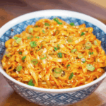 A blue patterned bowl filled with peanut chili oil noodles, topped with scallions and sesame seeds. The glossy sauce and chewy noodles make this dish a perfect weeknight fix.