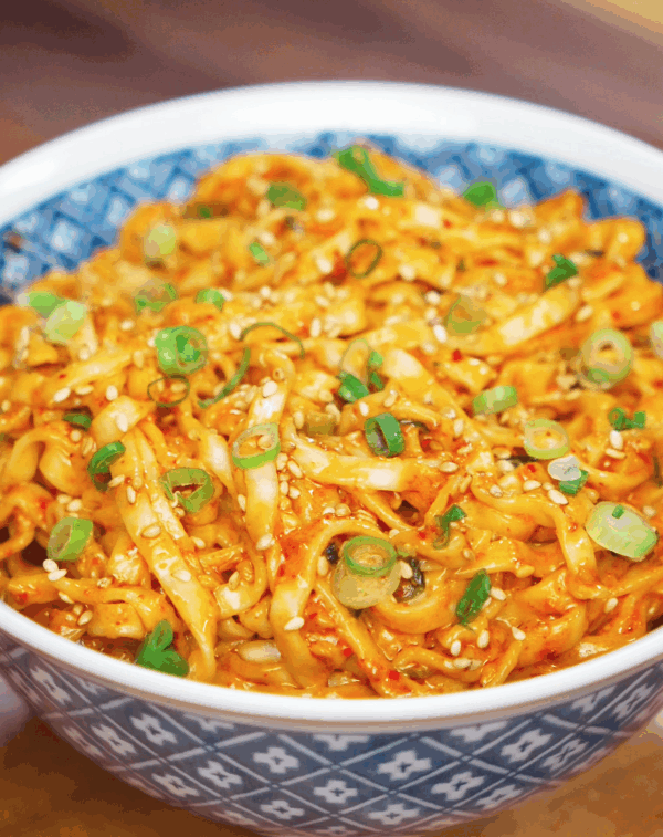 A blue patterned bowl filled with peanut chili oil noodles, topped with scallions and sesame seeds. The glossy sauce and chewy noodles make this dish a perfect weeknight fix.