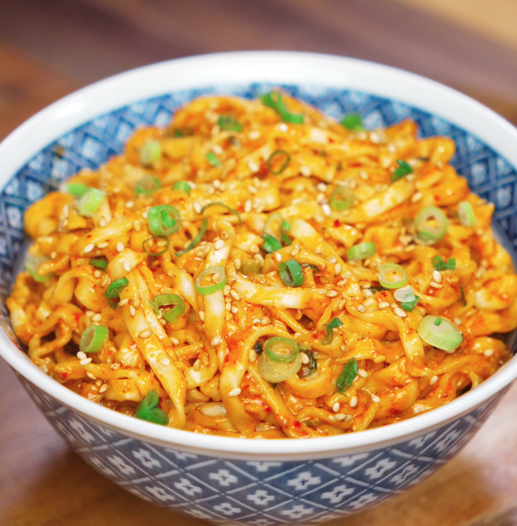 A blue patterned bowl filled with peanut chili oil noodles, topped with scallions and sesame seeds. The glossy sauce and chewy noodles make this dish a perfect weeknight fix.