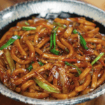 A bowl of glossy teriyaki udon noodles mixed with sautéed cabbage, carrots, garlic, and scallions, coated in a rich, savory-sweet sauce.