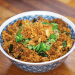 A patterned blue bowl filled with Thai basil fried rice, topped with golden fried onions and fresh scallions. The rice is coated in a rich, savory sauce and speckled with wilted Thai basil.