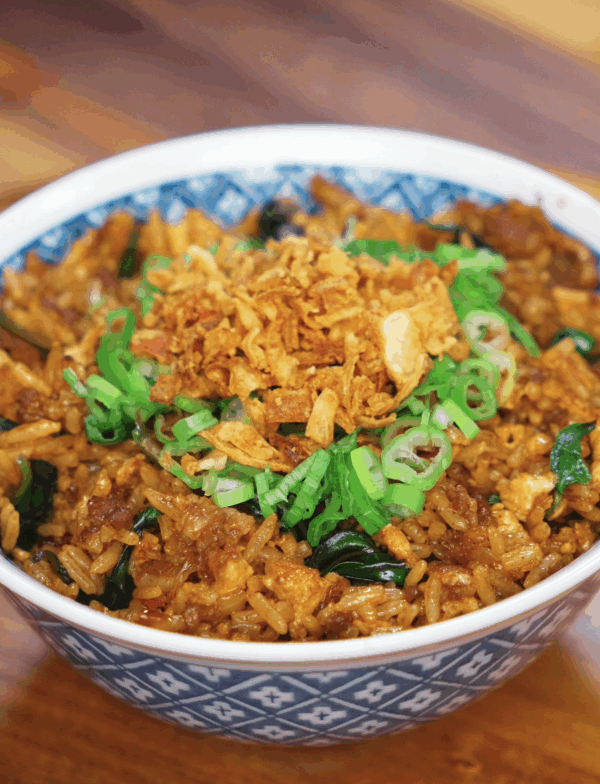 A patterned blue bowl filled with Thai basil fried rice, topped with golden fried onions and fresh scallions. The rice is coated in a rich, savory sauce and speckled with wilted Thai basil.