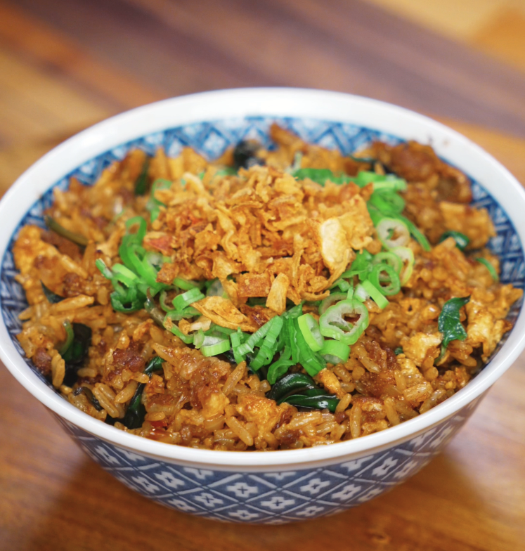 A patterned blue bowl filled with Thai basil fried rice, topped with golden fried onions and fresh scallions. The rice is coated in a rich, savory sauce and speckled with wilted Thai basil.