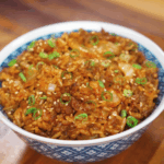 A bowl of spicy kimchi beef fried rice is garnished with scallions and sesame seeds, glistening with sauce and packed with texture.
