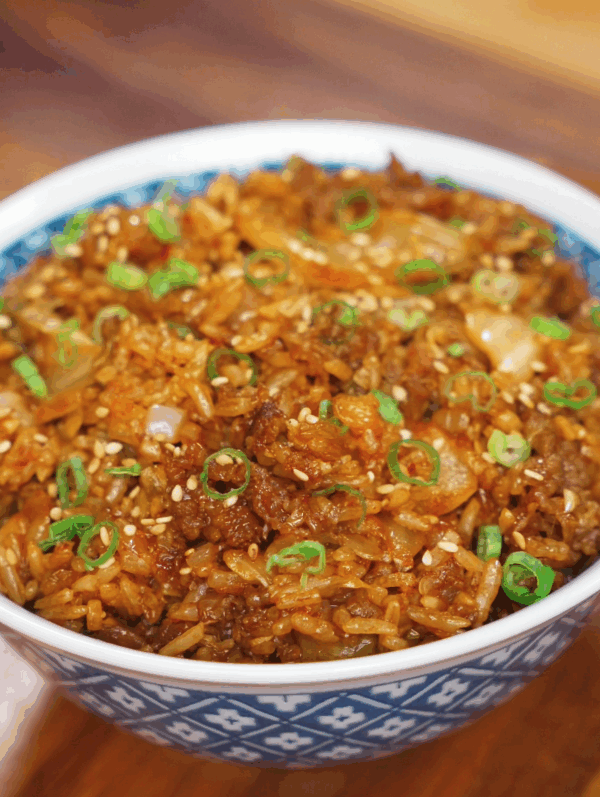 A bowl of spicy kimchi beef fried rice is garnished with scallions and sesame seeds, glistening with sauce and packed with texture.