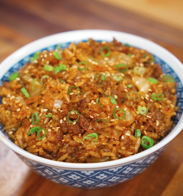 A bowl of spicy kimchi beef fried rice is garnished with scallions and sesame seeds, glistening with sauce and packed with texture.