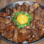A finished rice bowl with caramelized pork belly bites arranged in a circle, garnished with scallions and a raw egg yolk in the center for added richness.