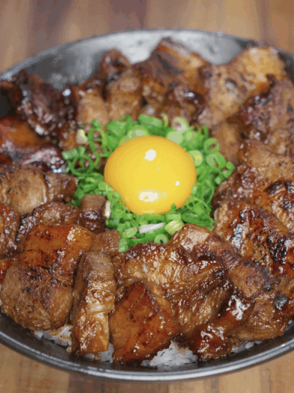 A finished rice bowl with caramelized pork belly bites arranged in a circle, garnished with scallions and a raw egg yolk in the center for added richness.