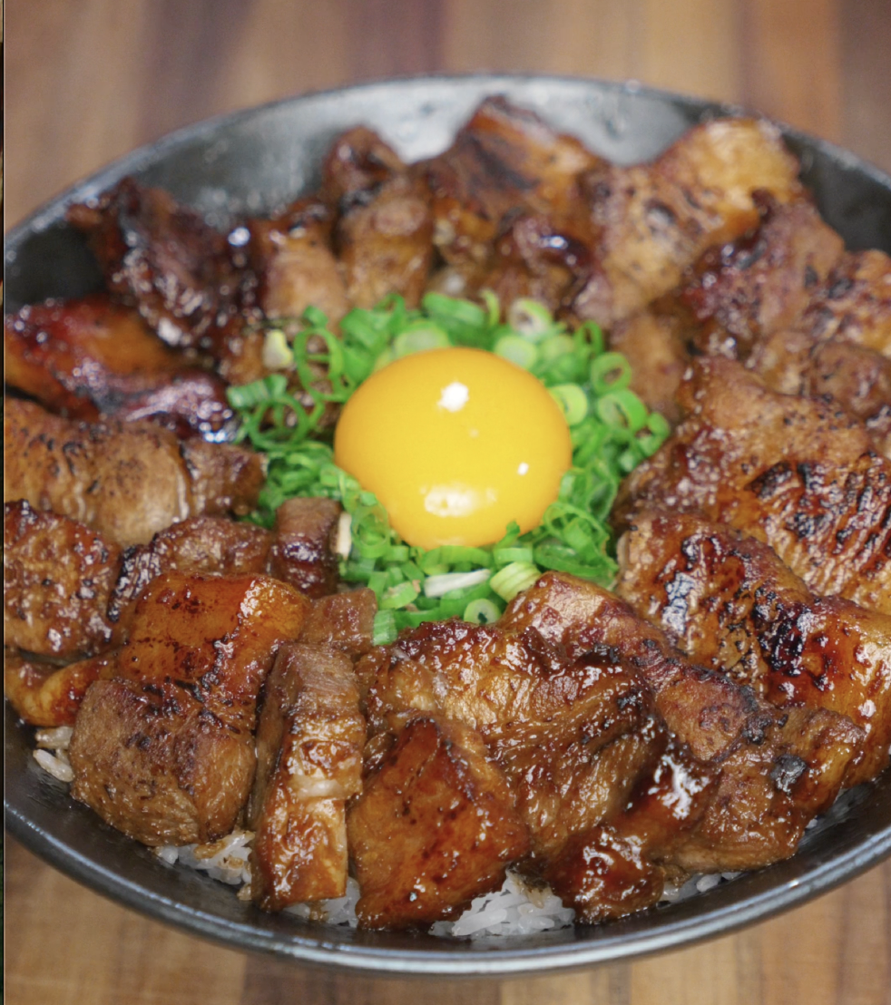 A finished rice bowl with caramelized pork belly bites arranged in a circle, garnished with scallions and a raw egg yolk in the center for added richness.