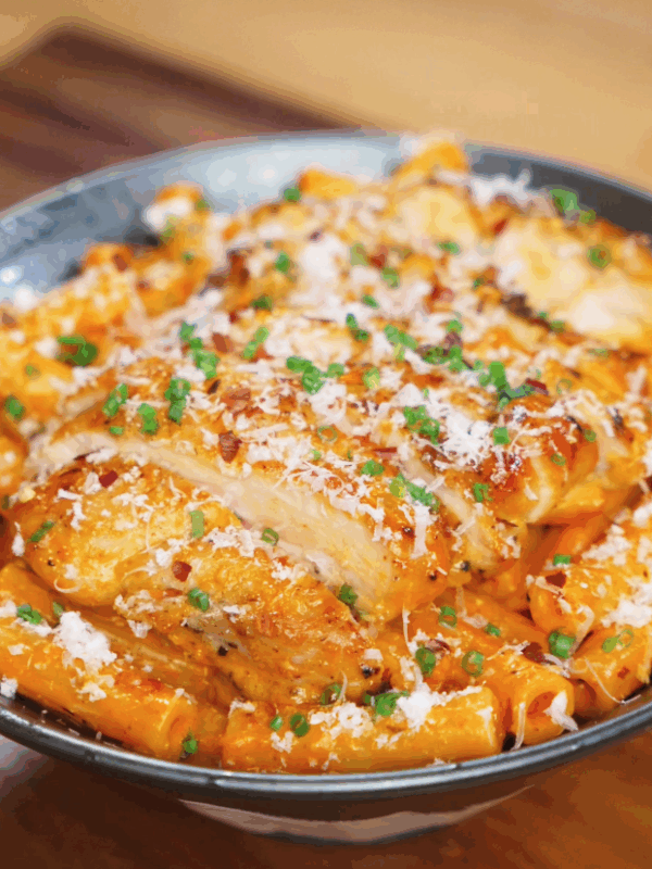 A generous bowl of rigatoni coated in creamy orange sauce is topped with juicy sliced chicken, fresh chives, chili flakes, and a shower of parmesan.