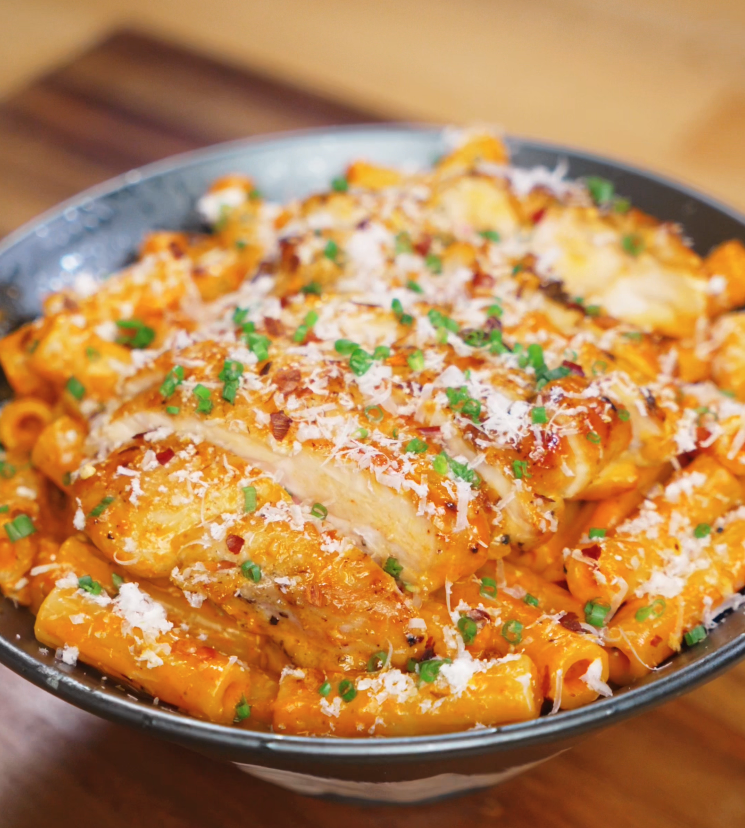 A generous bowl of rigatoni coated in creamy orange sauce is topped with juicy sliced chicken, fresh chives, chili flakes, and a shower of parmesan.