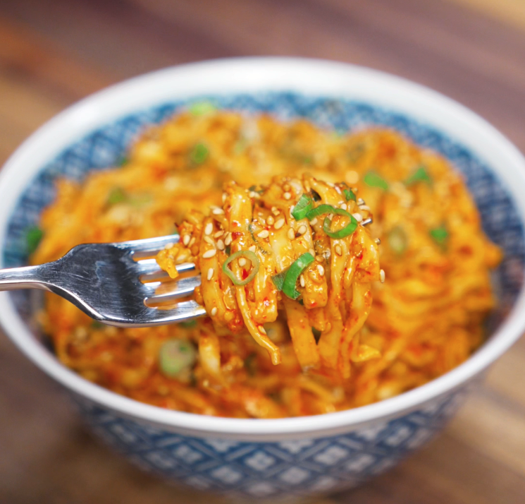 A fork lifts a glossy bite of peanut chili oil noodles from the bowl. The sauce clings to every noodle, garnished with green onions and sesame seeds for extra crunch.