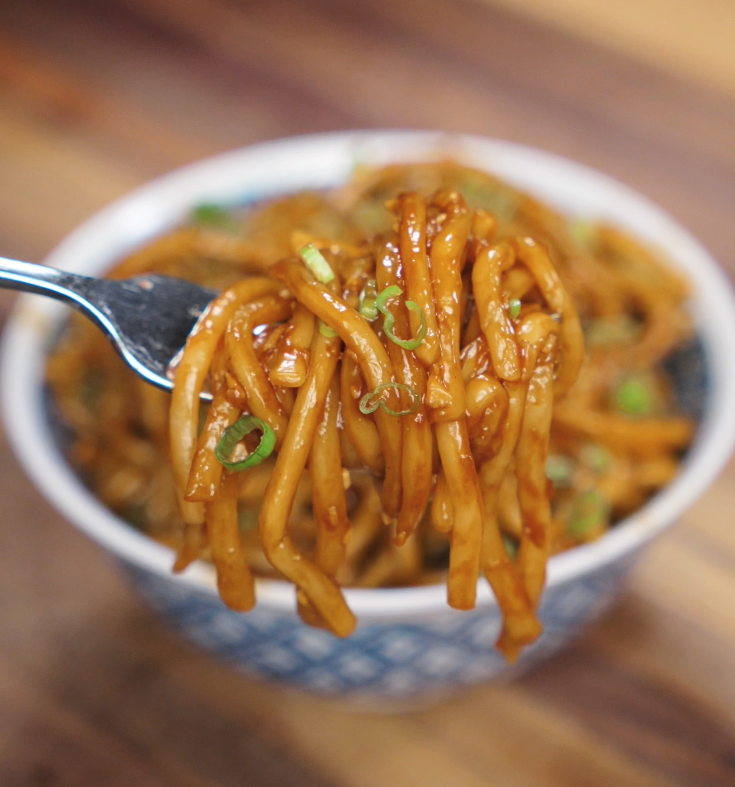 A close-up of a fork lifting tangled garlic noodles, showcasing their glossy, savory coating and fresh scallion garnish.