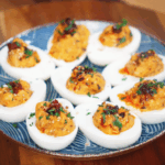 A plate of deviled eggs sits ready to serve, each filled with creamy chili crunch filling and finished with sesame seeds and herbs.