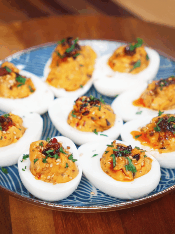 A plate of deviled eggs sits ready to serve, each filled with creamy chili crunch filling and finished with sesame seeds and herbs.