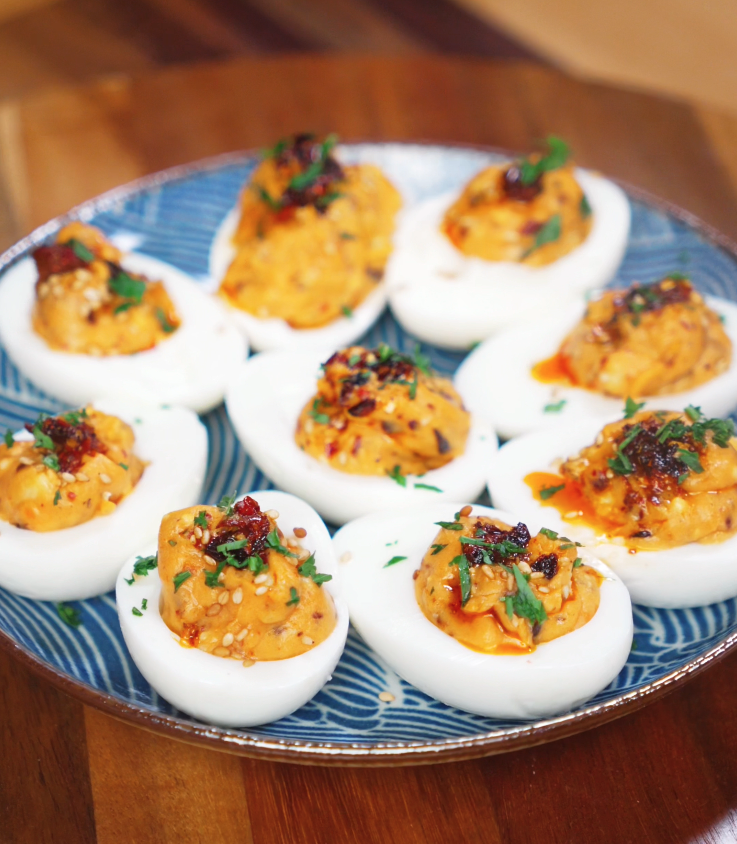 A plate of deviled eggs sits ready to serve, each filled with creamy chili crunch filling and finished with sesame seeds and herbs.