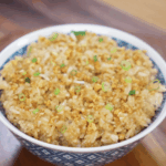 A full bowl of garlic fried rice sits on a wooden table, topped with crispy garlic bits and freshly sliced scallions for garnish.