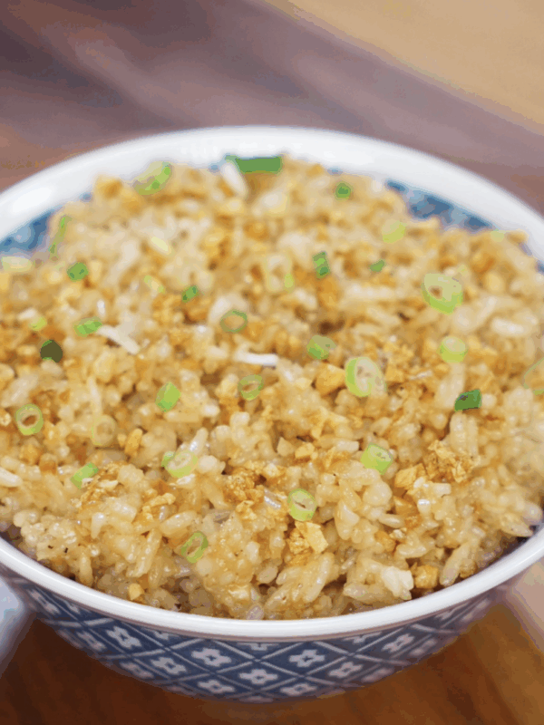 A full bowl of garlic fried rice sits on a wooden table, topped with crispy garlic bits and freshly sliced scallions for garnish.