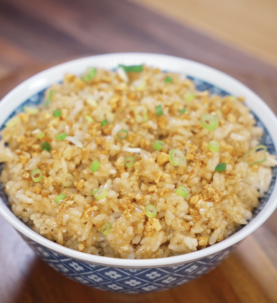 A full bowl of garlic fried rice sits on a wooden table, topped with crispy garlic bits and freshly sliced scallions for garnish.