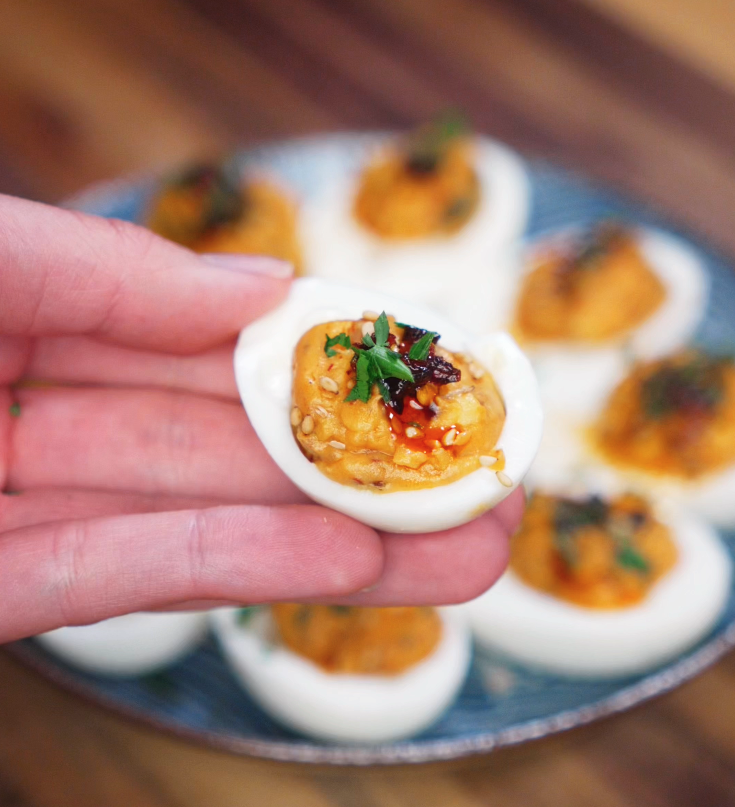 A single chili crunch deviled egg is held up to the camera, showing the creamy yolk center topped with chili oil, sesame seeds, and chopped parsley.