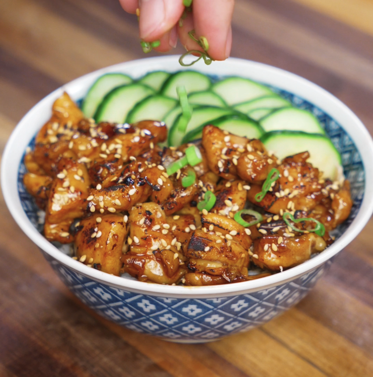 A hand sprinkles freshly sliced scallions over a bowl of garlic butter chicken, featuring golden, caramelized chicken glazed in a savory garlic sauce, topped with sesame seeds and served with sliced cucumber over rice.