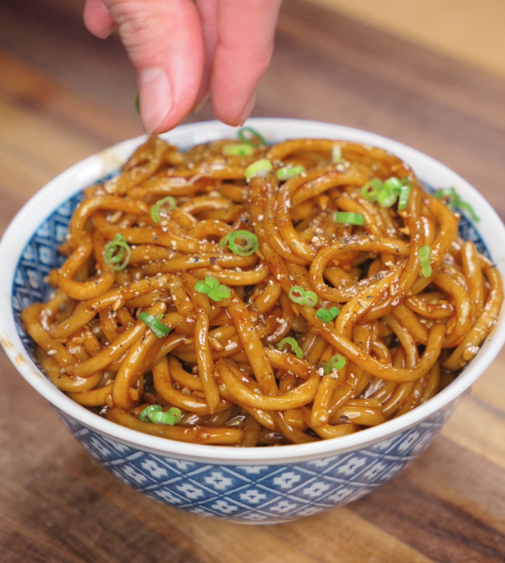 A hand sprinkles sliced scallions over the bowl of garlic noodles, adding a pop of freshness and color.