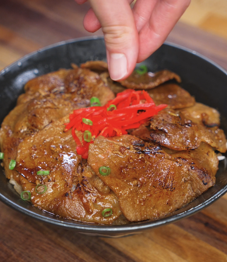 A hand sprinkles vibrant red pickled ginger and scallions over a bowl of glazed pork, finishing off the dish with color and freshness.