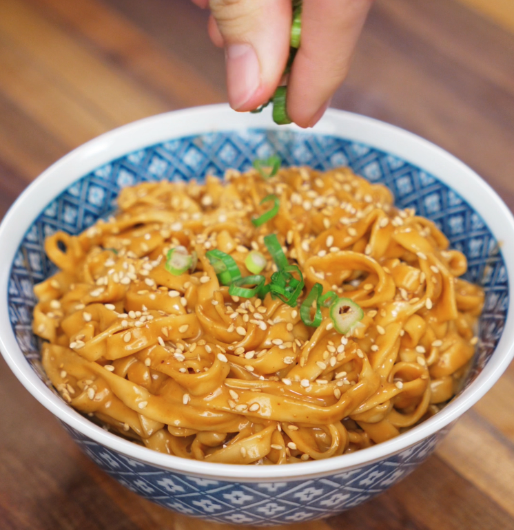 A hand finishes the dish by sprinkling fresh scallions and sesame seeds over the creamy sesame peanut noodles for crunch and color.