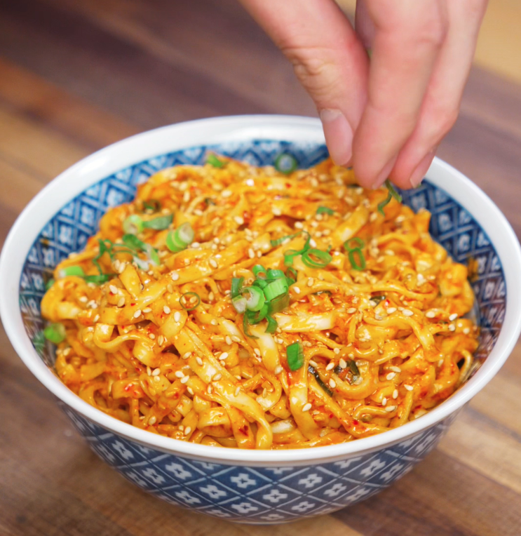 A hand sprinkles fresh green scallions over a bowl of peanut chili oil noodles. The vibrant garnish pops against the spicy orange noodles.