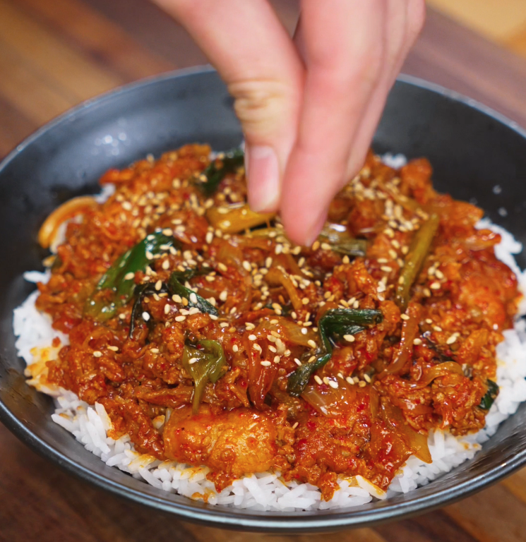 A hand sprinkles toasted sesame seeds over a freshly plated bowl of pork bulgogi, adding texture and a nutty finish to the dish.