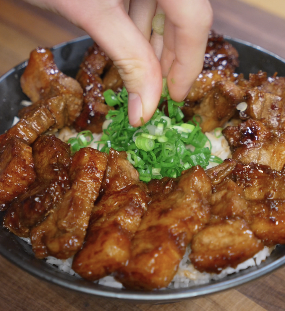 A hand sprinkles chopped scallions over a glossy bowl of sticky pork belly bites served on rice. The caramelized glaze and green onions add a vibrant finish to the rich dish.