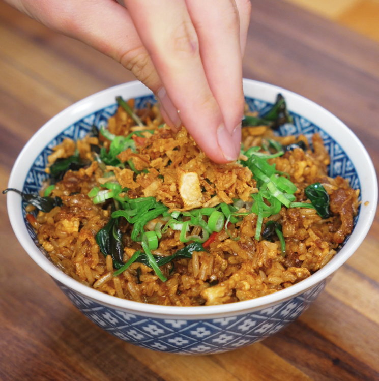 A hand sprinkles crispy fried onions over a bowl of Thai basil fried rice, topped with fresh scallions and filled with saucy beef and wilted basil leaves.