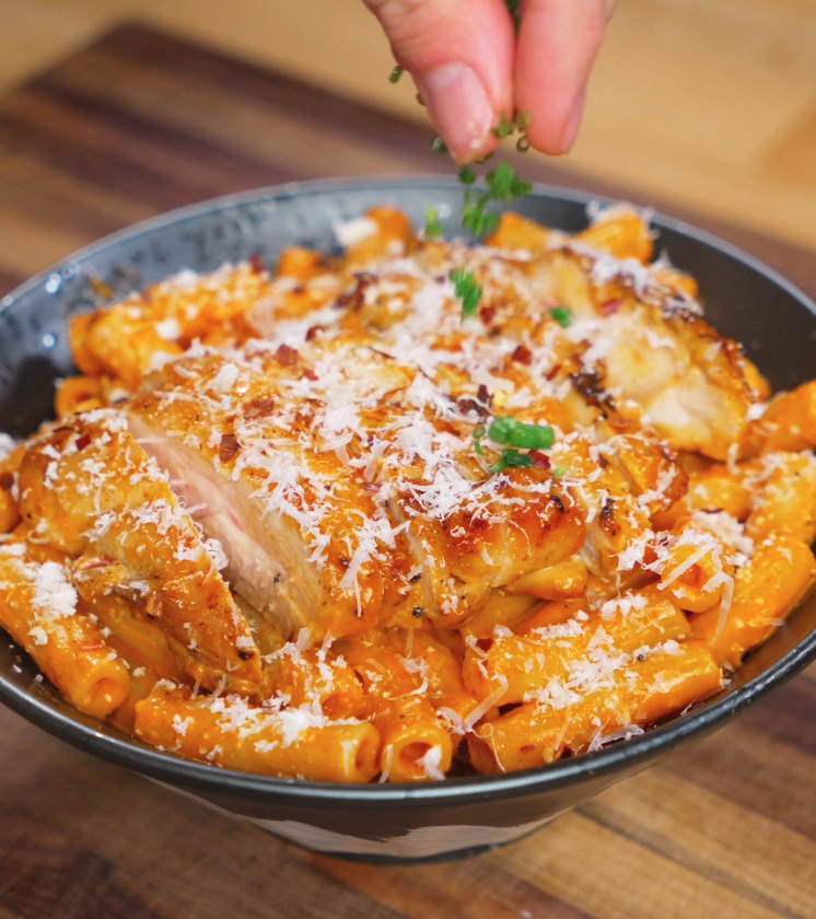 A hand sprinkles chopped chives over a bowl of spicy creamy chicken pasta, topping the rigatoni and glazed chicken with fresh green color and a final hit of flavor.