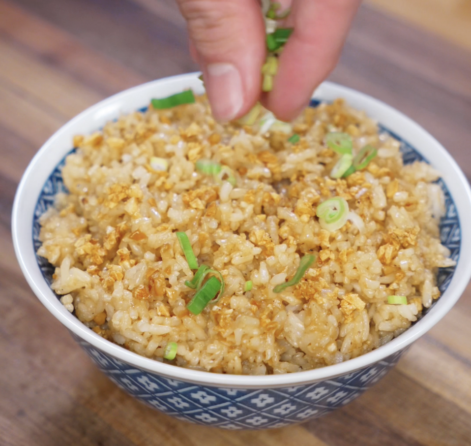 A hand sprinkles fresh scallions over a bowl of garlic fried rice, adding a pop of color and freshness to the golden dish.