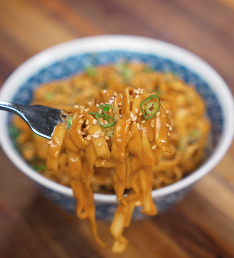 A fork lifts a twirl of saucy sesame peanut noodles, showing the creamy coating and garnishes of scallions and sesame seeds.